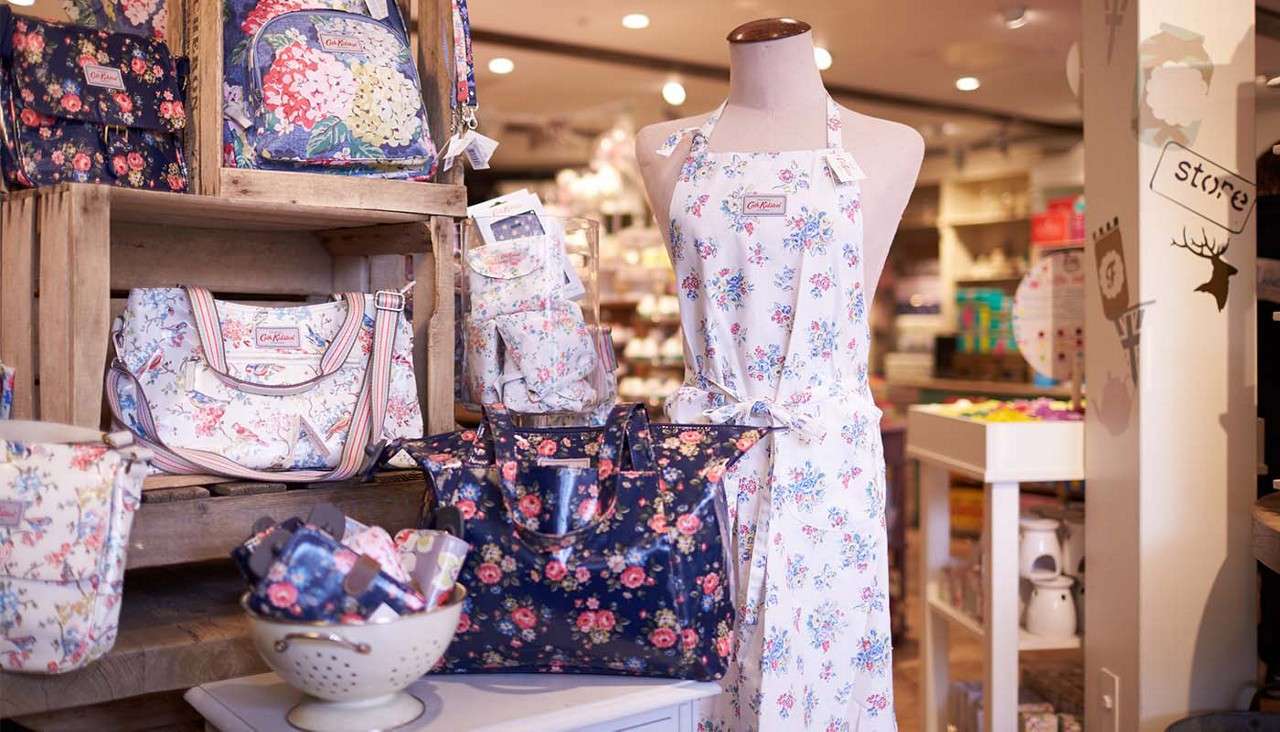 Mannequin wearing a white floral apron stands amid matching floral handbags, pouches, and a bowl of small items on wooden crates in a boutique. Text: “Cath Kidston” (labels), “store” (wall sign).