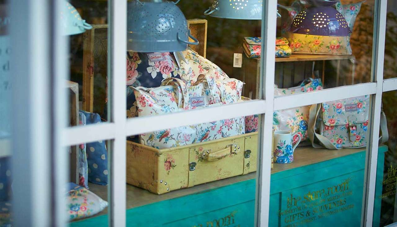 Floral handbags and a mug sit displayed on shelves and a vintage suitcase, illuminated by colander-shaped lamps, behind a shop window on a teal counter.

Text: “Cath Kidston” (repeated on bag labels); “the store room”; “GIFTS & SOUVENIRS”; [additional small gold text illegible].