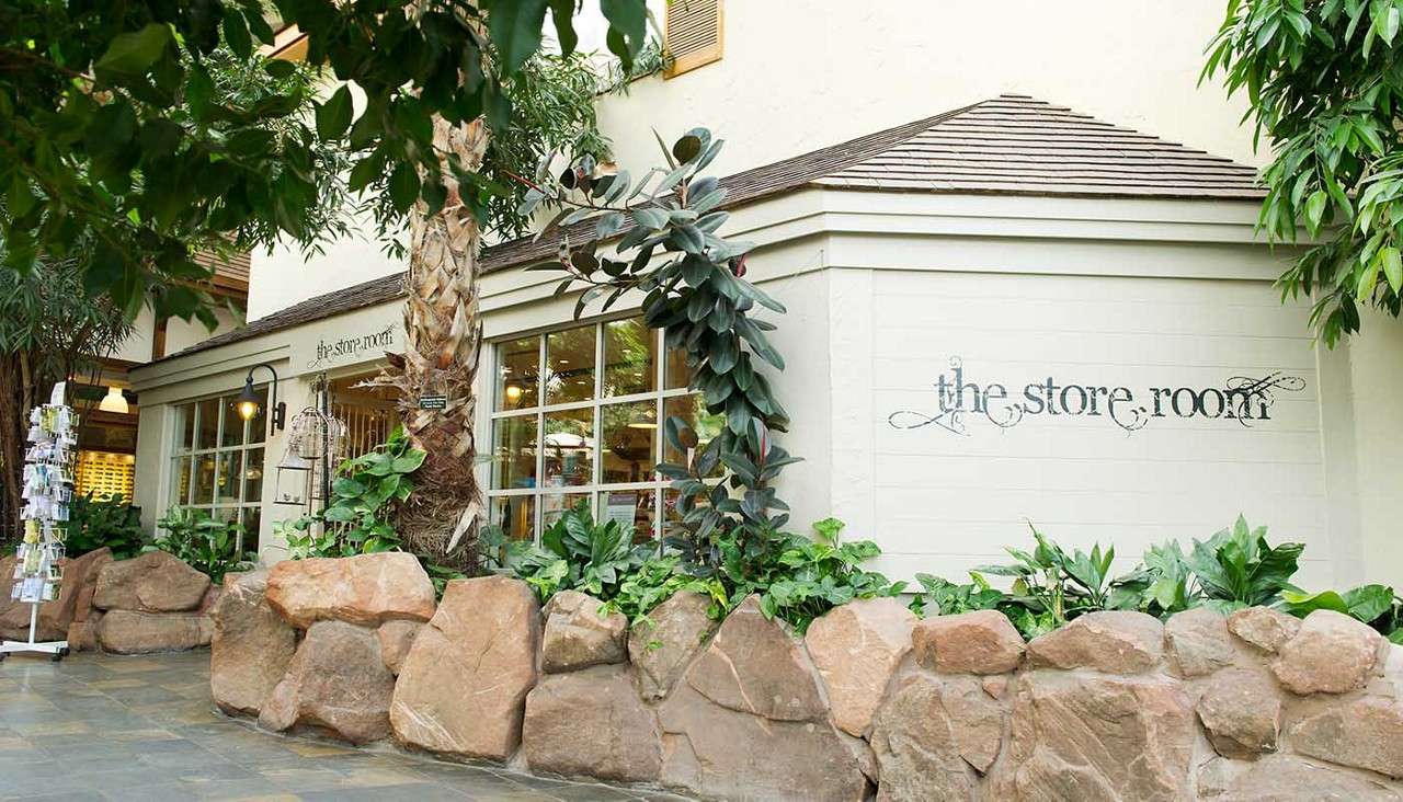 Storefront displays goods through large windows, framed by rock planters and lush indoor plants in an atrium. Sign reads: "the store room." A postcard rack stands nearby under warm lights.