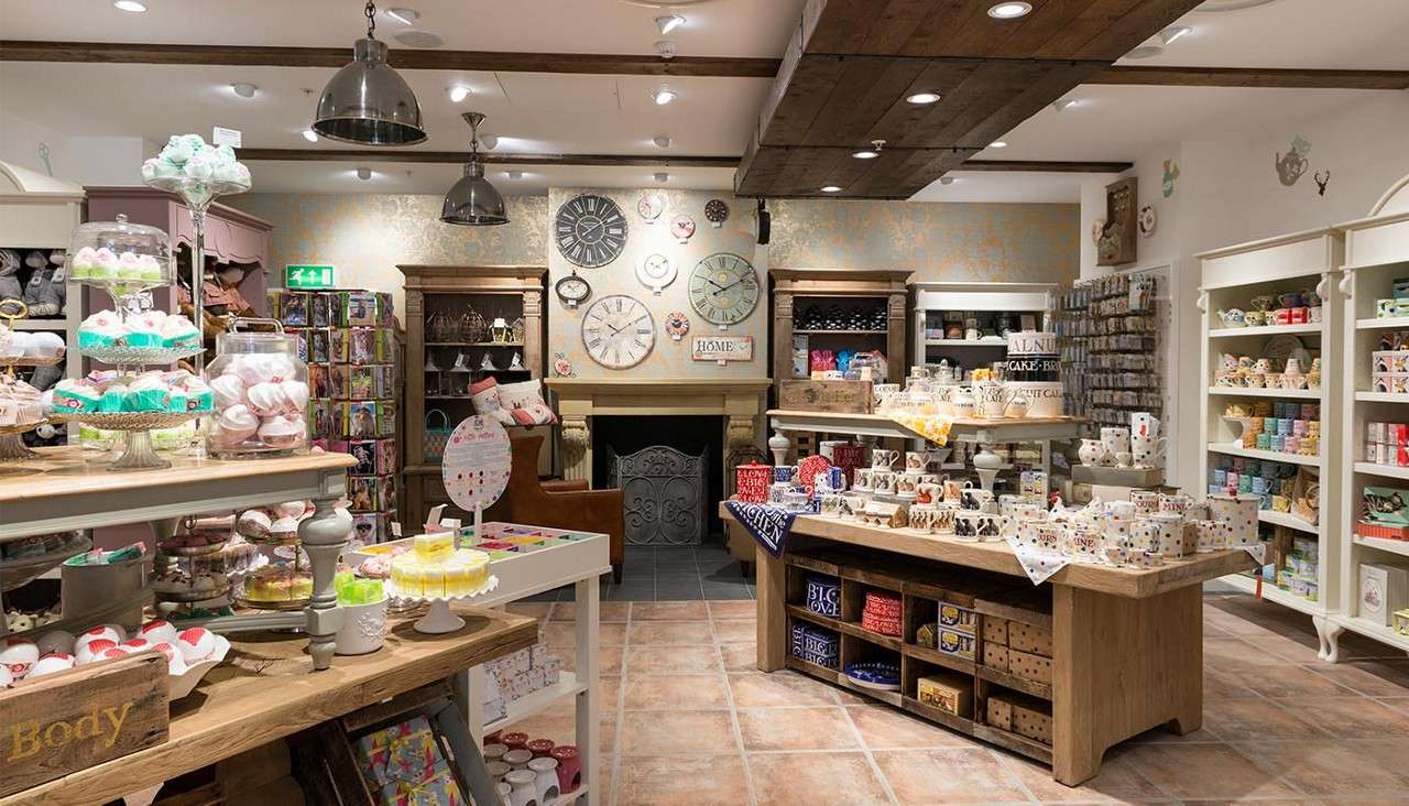 Shelves of mugs and gifts—arranged and displayed—fill a cozy boutique interior with wooden tables, an empty fireplace, pendant lights, and wall clocks. Text: HOME; BIG LOVE; Body.