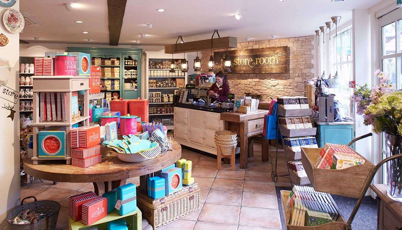Shop clerk rings up purchases at a rustic boutique counter, surrounded by colorful gift displays and shelves of goods under warm lights. Visible sign: "store room".