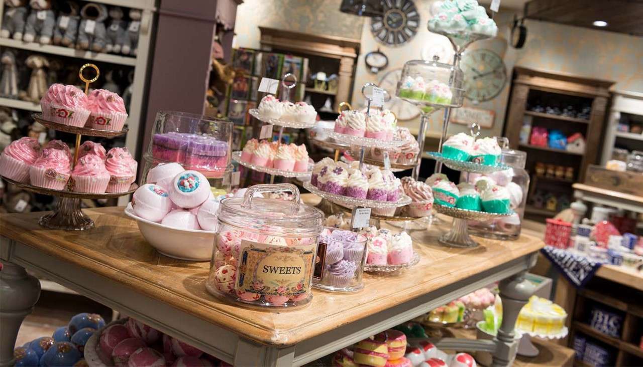 Cupcake-shaped items are displayed on tiered trays, clustering on a wooden table; shelves and decor surround the boutique-like shop. Text on a jar reads: SWEETS.