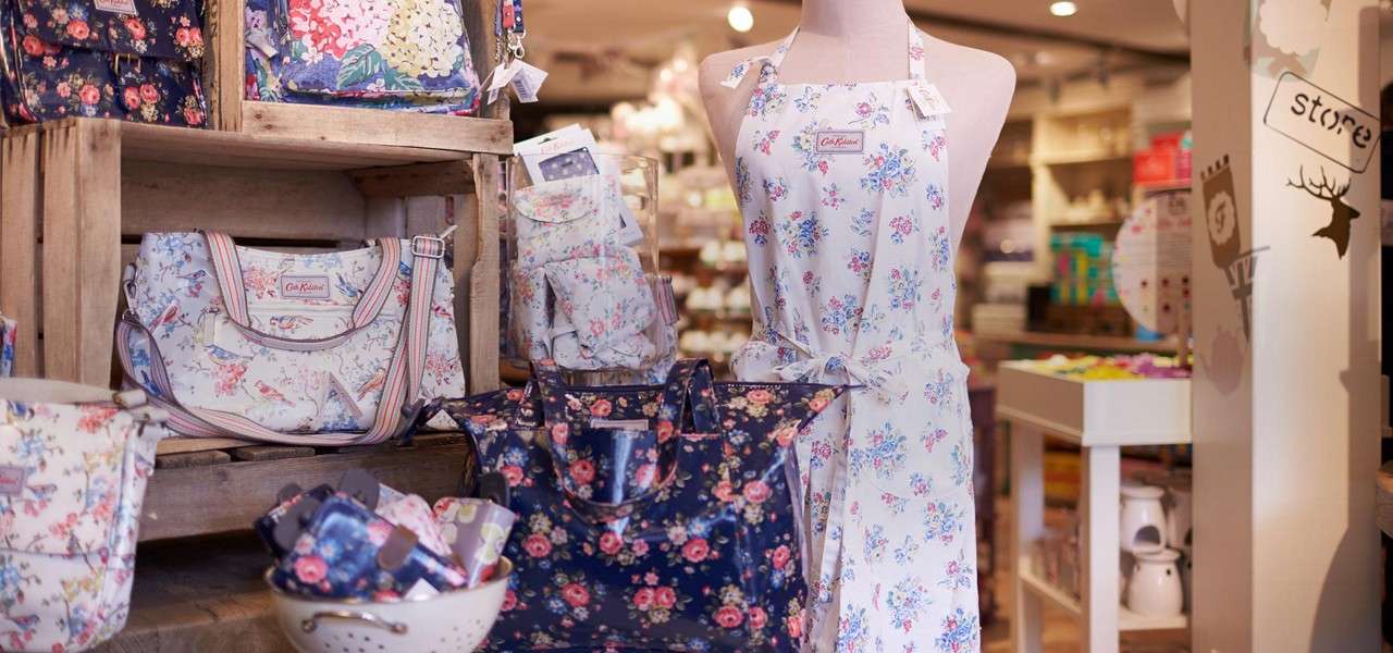 Mannequin wearing a floral apron stands among matching floral bags and accessories displayed on wooden crates in a boutique. Text: “Cath Kidston”; “store”.