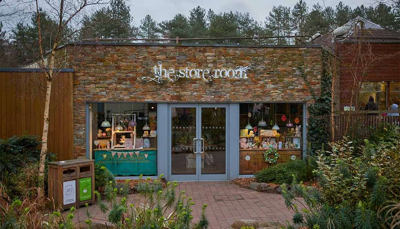 Shopfront displays gifts in lit windows, seen through glass double doors; context: stone facade, landscaped shrubs, bin, wooden building and trees behind. Text visible: "the store room".