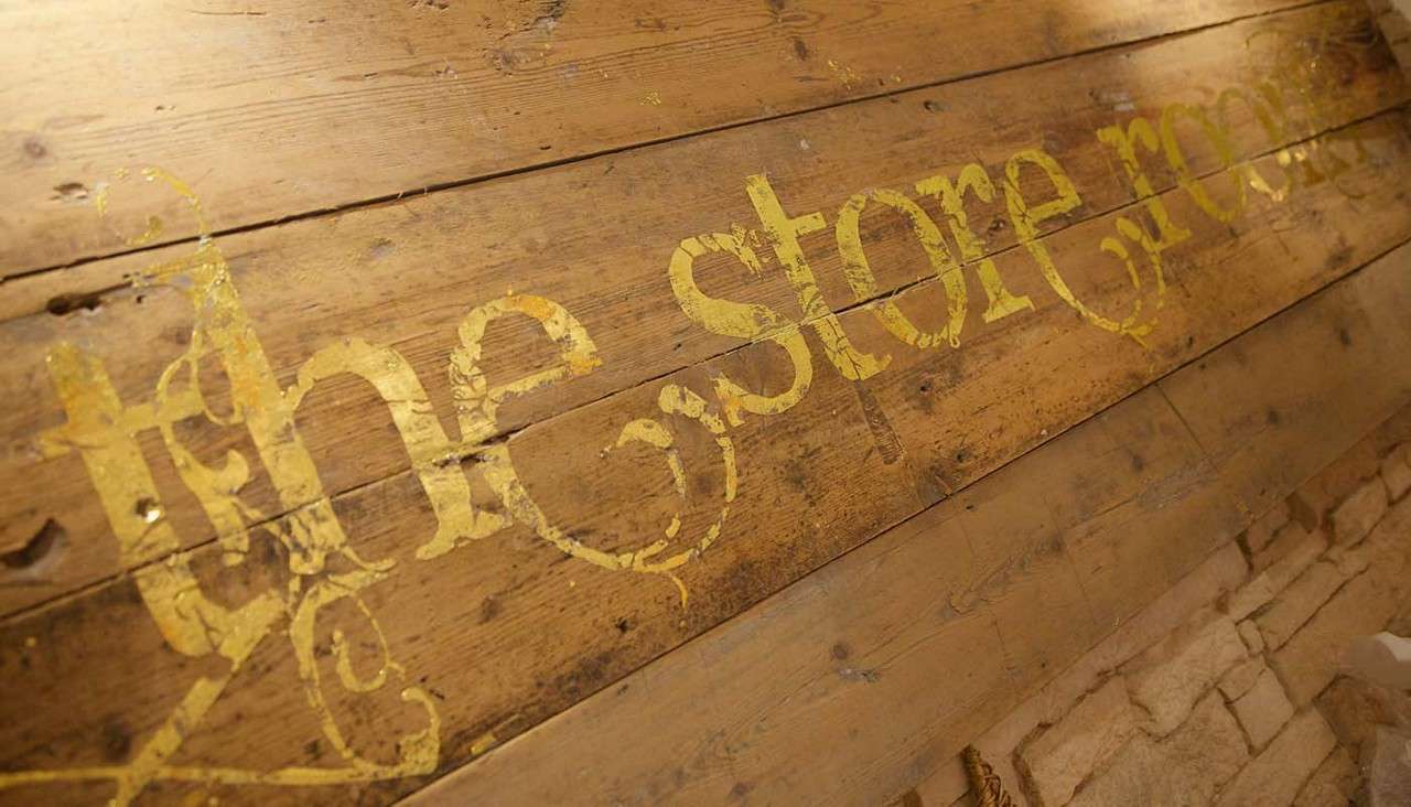 Gold, weathered lettering reads “the store room,” stenciled across aged wooden planks at a diagonal; context shows a rustic interior with a stacked stone wall along the bottom edge.