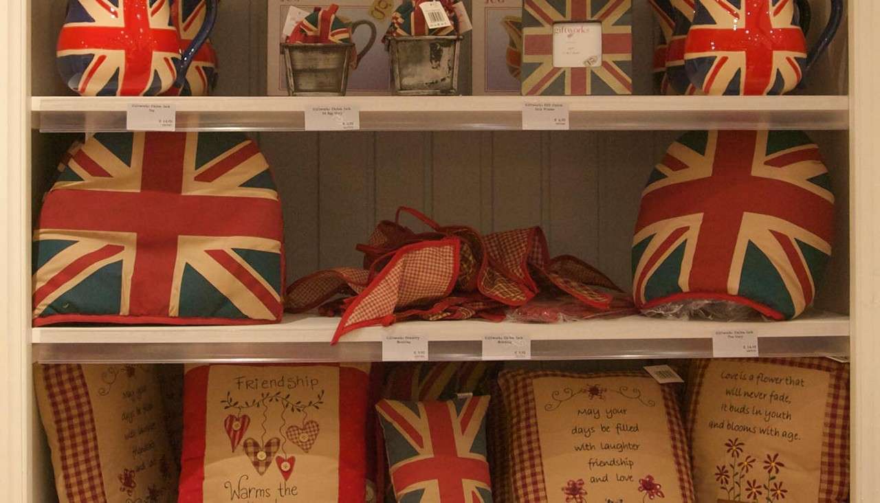 Union Jack–patterned pitchers, tea cozies, and cushions sit neatly displayed on shop shelves.
Text: “Friendship Warms the”
“may your days be filled with laughter friendship and love”
“Love is a flower that will never fade, it buds in youth and blooms with age”
“giftworks”