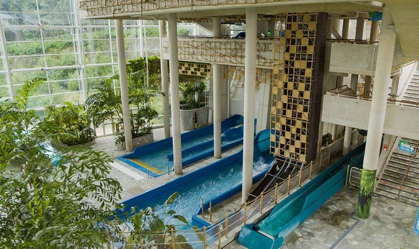 Blue water slide channels carry churning water, twisting through an indoor aquapark; white columns, glass walls, tropical plants, and stairways surround the flumes in a bright, greenhouse-like atrium.