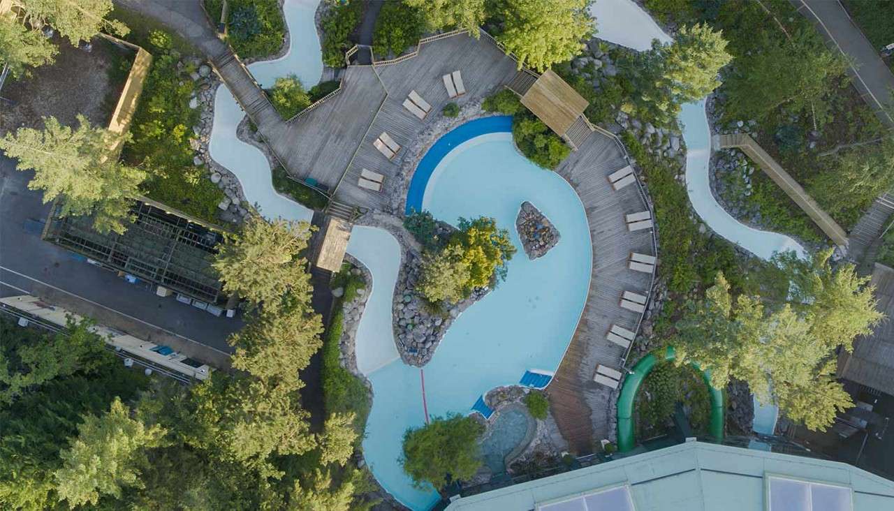 Curving outdoor pool sits empty; lounge chairs line wooden decks while a lazy river winds through landscaped trees and rocks in a water-park setting.