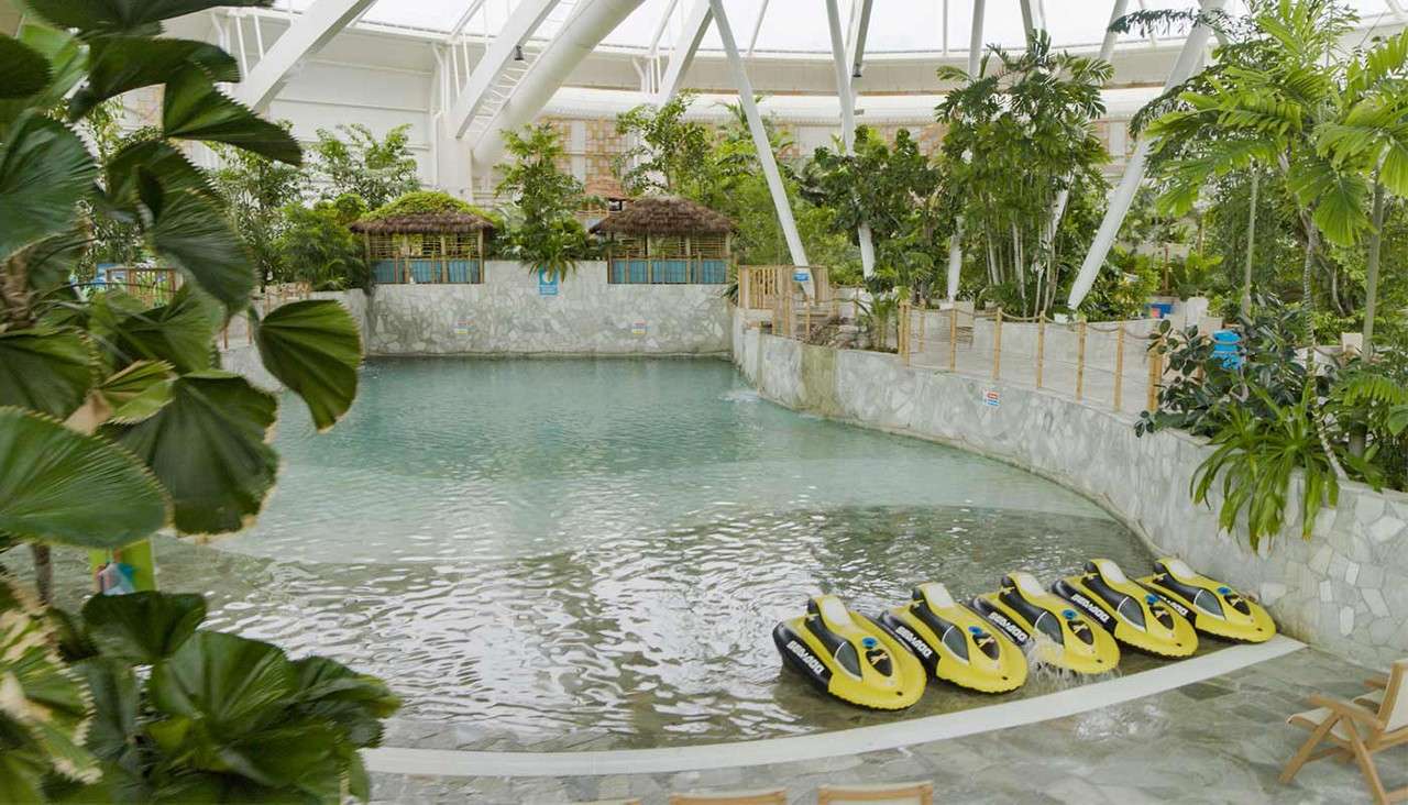 Yellow water scooters sit lined up at a shallow pool edge, within an indoor tropical water park, framed by palm-like plants, stone walls, and thatched cabanas under a skylit roof.