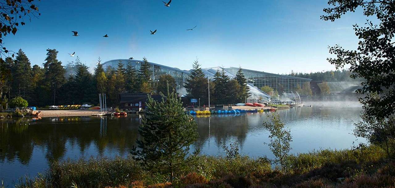Lakeside resort—kayaks rest on shore; birds glide overhead; glass-domed buildings gleam behind pine trees; morning mist lifts over calm water in a forested park setting.