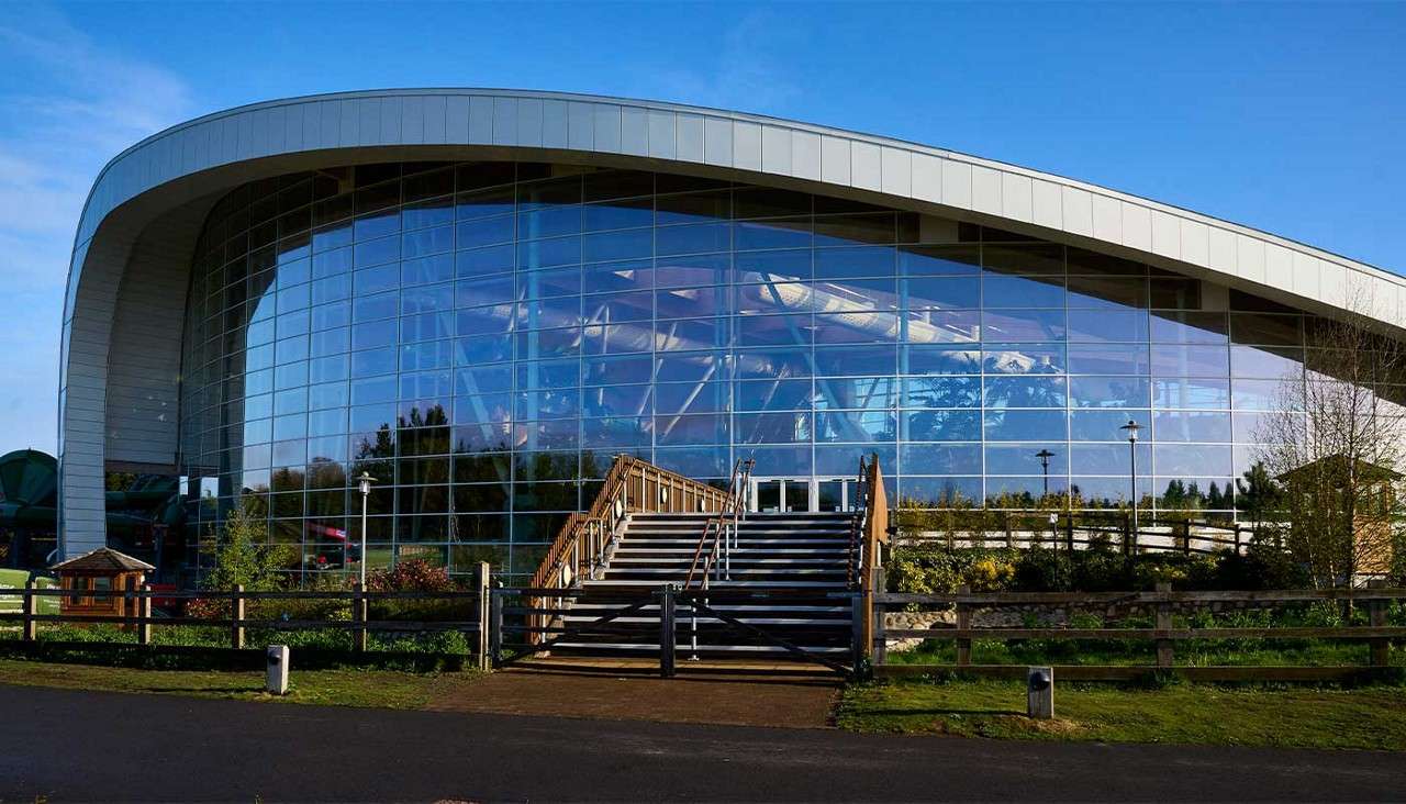 Curved-roof glass building reflects blue sky and trees; wide central staircase leads to main entrance. Landscaped shrubs, wooden fence, and lampposts surround it on a sunny day.