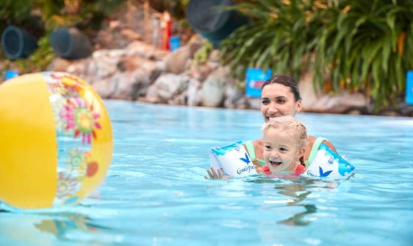Child wearing “Center Parcs” armbands swims toward a yellow floral beach ball, guided by an adult in a pool, with surrounding rocks, lush plants, and poolside features.