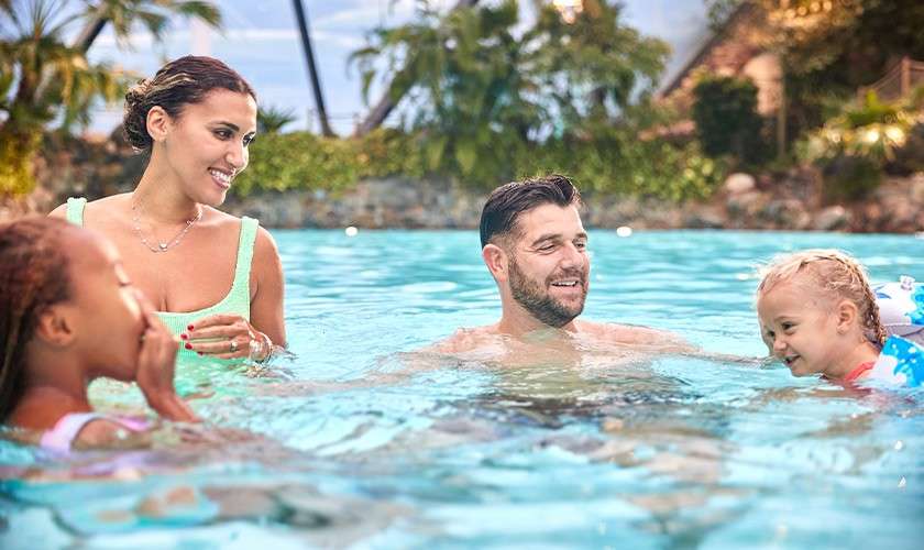 Family of four swims and smiles, interacting playfully in a clear blue pool; adults watch children splashing with floaties; palm trees, rocks, and resort buildings surround the tropical outdoor setting.