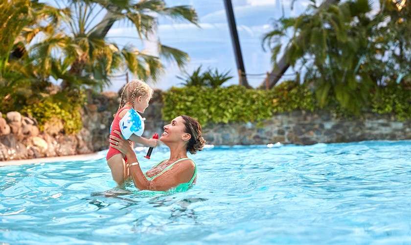 Woman lifts a toddler in floaties while standing in a bright blue pool; both smiling. Tropical plants and a stone pool edge surround them under daylight.