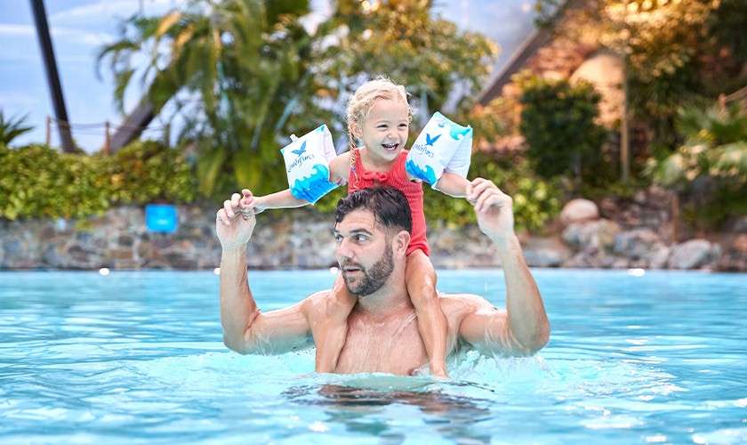 Child wearing blue arm floaties rides on an adult’s shoulders, both smiling and splashing, in a bright outdoor swimming pool surrounded by tropical plants, rocks, and resort-style buildings.