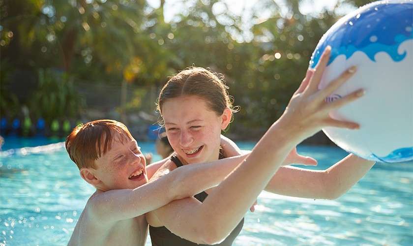 Two children hug and laugh while reaching for a beach ball in a sunlit outdoor swimming pool, surrounded by tropical plants and trees.