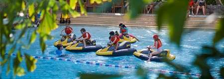 Inflatable bumper boats bump and circle as riders paddle in a swimming pool, while a guide in the water supervises; leafy branches frame the scene near poolside seating.