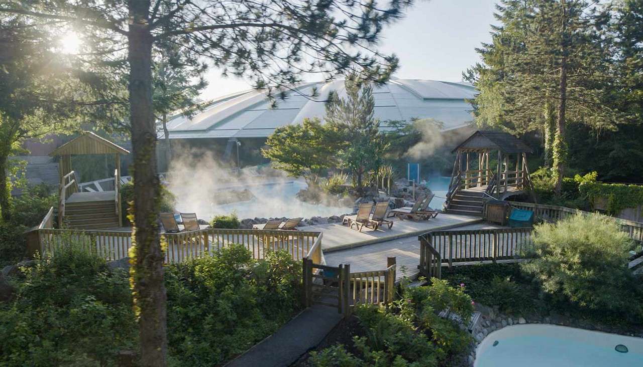 Steaming outdoor pool sends mist upward, bordered by wooden decks and lounge chairs, with small footbridges. Set amid lush trees and gardens, a large domed building rises in the background.