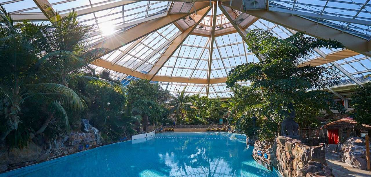 Large indoor swimming pool reflects sunlight, lying still. Surrounded by tropical plants, rock features, and a statue beneath a vast glass-domed roof, suggesting a serene, climate-controlled waterpark.