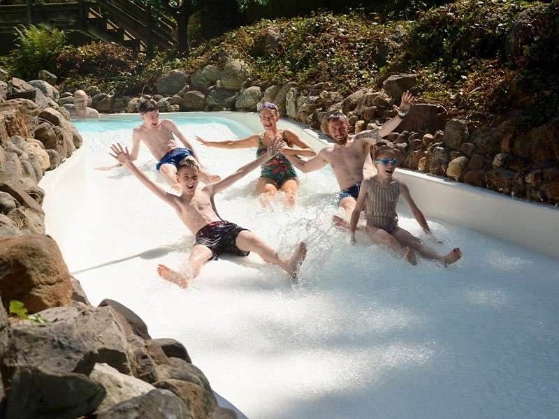 Group of people slide down a shallow water chute, splashing and raising arms, in a rock-lined outdoor setting with sunlight and foliage, while another person follows behind in the channel.