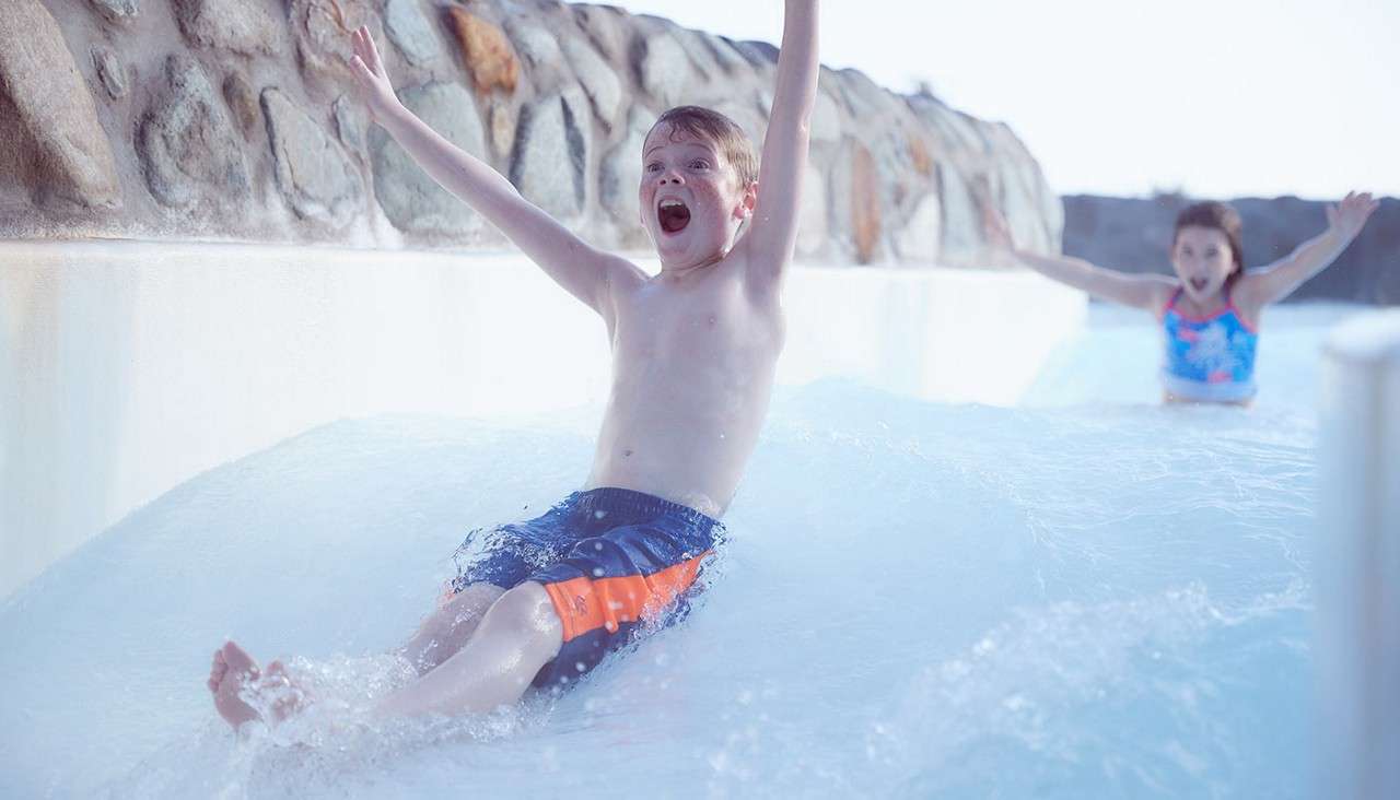 Boy raises arms, sliding feet-first down a water slide, splashing. Behind him, another child spreads arms. Context: outdoor waterpark channel bordered by a stone-faced wall under bright light.