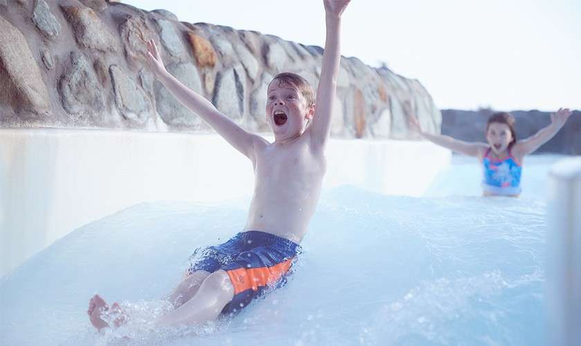 Child rides down a water slide, arms raised and mouth open, splashing into shallow water; another child follows behind; white slide runs alongside a stone wall in bright outdoor light.
