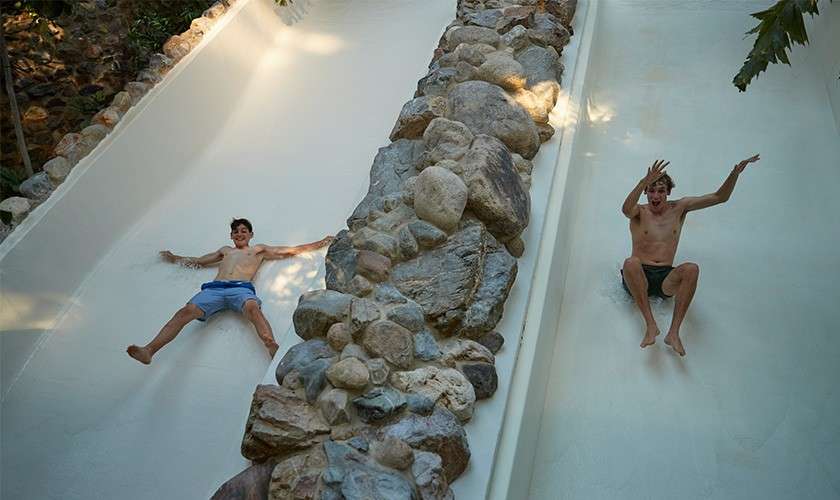 Two shirtless men slide separately down parallel white water slides, arms out, legs spread, separated by a rocky divider, under dappled sunlight with trees and stones around.