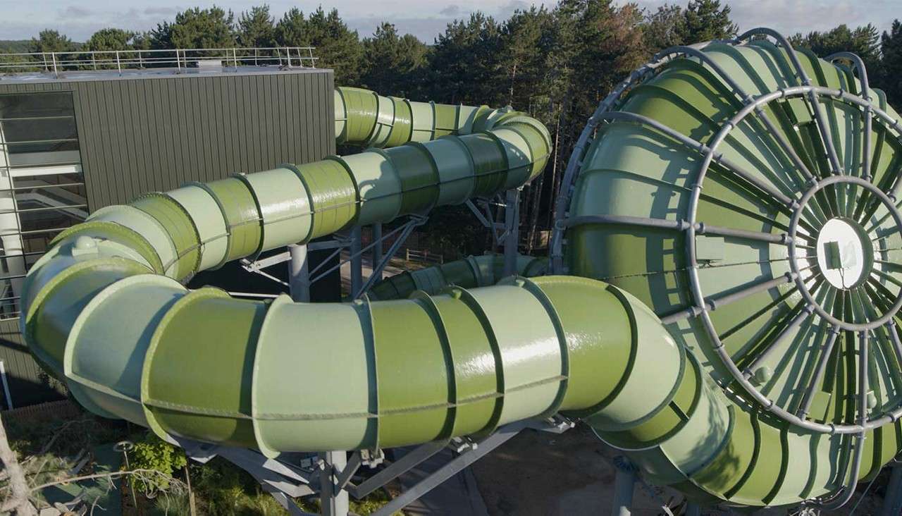 Enclosed green waterslide tubes twist overhead, feeding into a giant circular funnel structure; elevated supports beside a modern building, with surrounding trees and a clear sky.