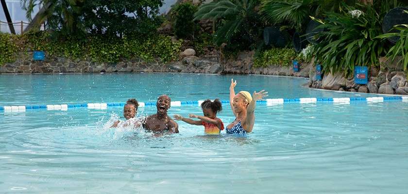 Family of four plays, splashing water, smiling and raising arms. They stand waist-deep in a large outdoor swimming pool, bordered by tropical plants, rocks, and floating lane markers.