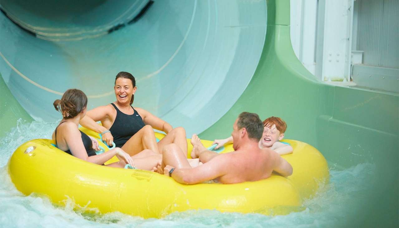 Four people ride a yellow inflatable raft, exiting a large enclosed water slide into a splash pool inside an indoor waterpark.