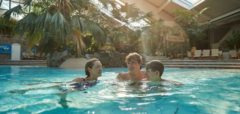 Three people swim and chat in a bright indoor pool, surrounded by palms and lounge chairs under skylights; a wooden sign in back reads “Venture Bay.”