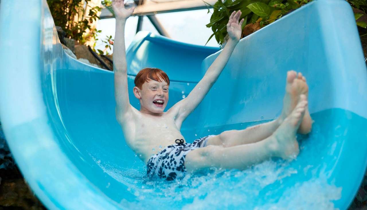Child raises arms while sliding feet-first down a curving blue water slide, splashing in shallow water, surrounded by plants and indoor structural beams.