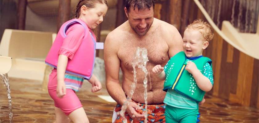 Adult man and two young children splash and laugh around a gushing floor fountain, wearing colorful life vests. Context: shallow splash pad inside an indoor water play area with slides.