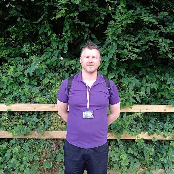 Man stands facing forward with hands behind his back, wearing a purple polo, backpack straps, and a lanyard badge, against a wooden fence and dense green foliage outdoors.