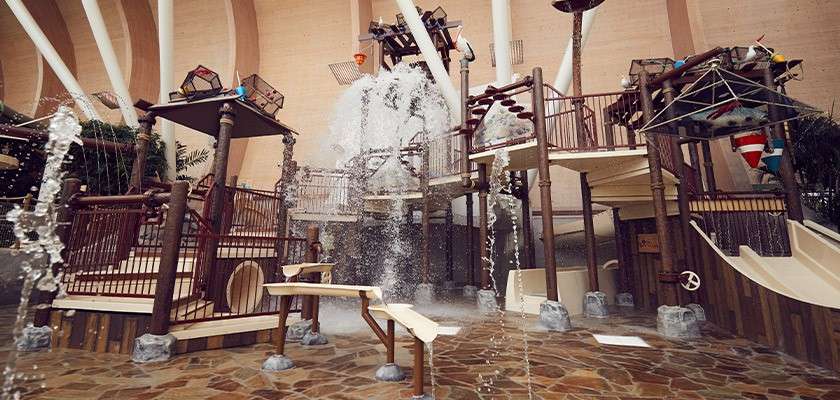 Multi-level water play structure splashes as a giant tipping bucket dumps water; fountains spray around bridges, stairs, and slides inside a large, arched-roof indoor aquatic park.