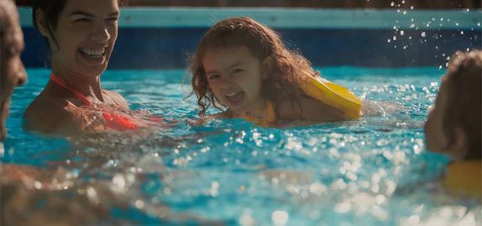 Child wearing yellow floaties laughs while paddling, supported by an adult, in a bright blue swimming pool; water splashes and two other people are partially visible nearby.