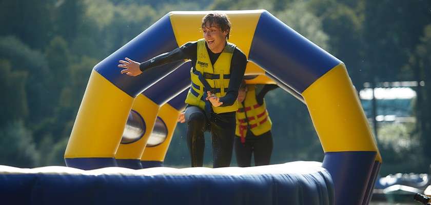 Smiling person runs and balances on a wet inflatable obstacle under blue-yellow arches, wearing a life vest and wetsuit, while another participant follows behind, in a sunny lakeside forest setting.