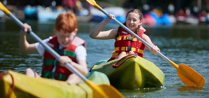 Two children paddle kayaks across a calm lake, wearing red life jackets and smiling, with other colorful boats blurred in the background under bright daylight.