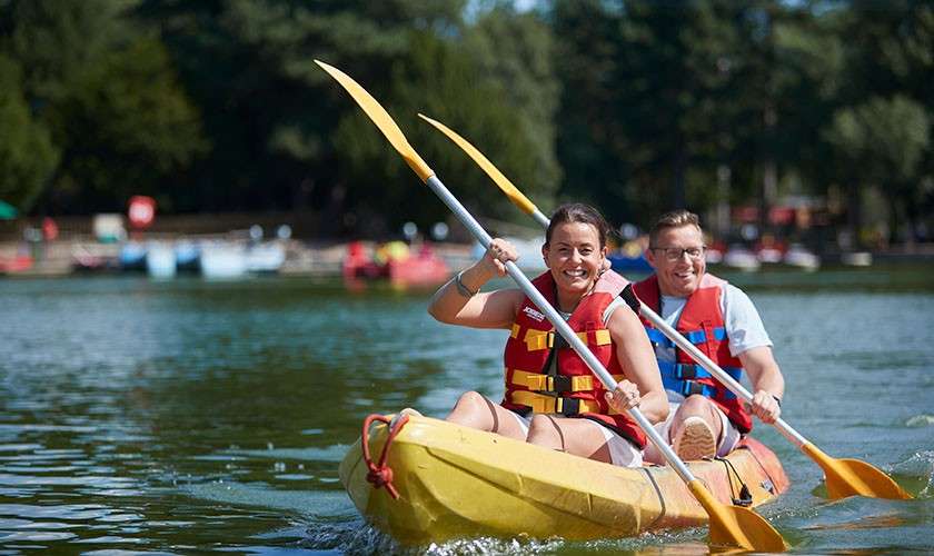 Two adults in red life vests paddle a yellow tandem kayak, water splashing at the bow, on a calm lake surrounded by trees and distant boats near a shoreline.