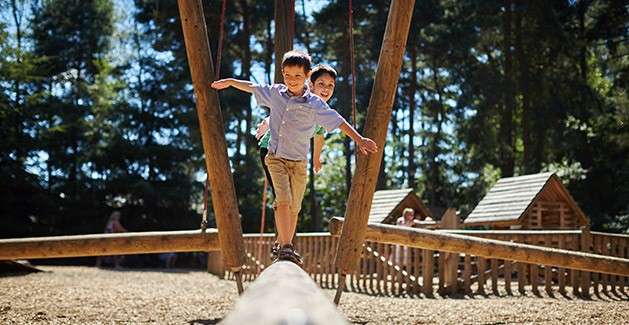 Two children balance with arms out while walking along a narrow wooden beam, smiling. Surroundings: outdoor playground with wooden structures, mulch ground, and tall trees under bright sunlight.