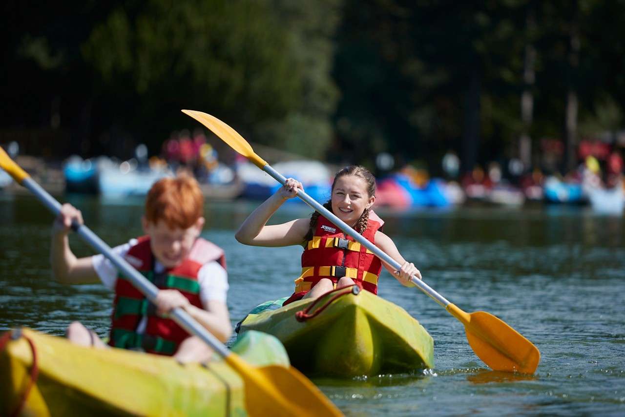 Children paddle kayaks on a calm lake, wearing red life jackets and using yellow paddles; background shows additional boats and trees. Text: JOBE.
