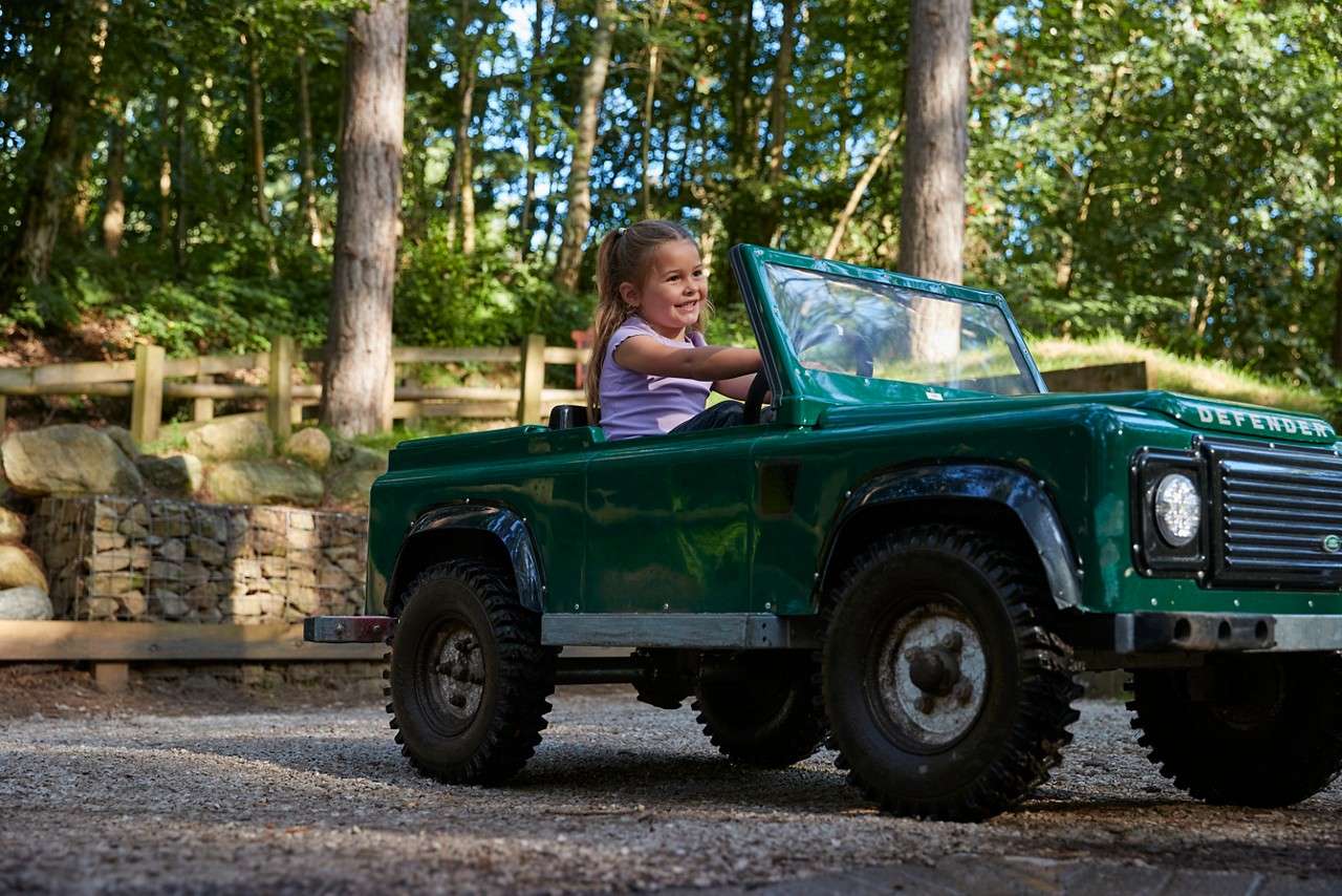 Child in a green miniature 4x4 drives, smiling; gravel path in a wooded park, with trees, a wooden fence, and a stone retaining wall. Text on hood: DEFENDER.
