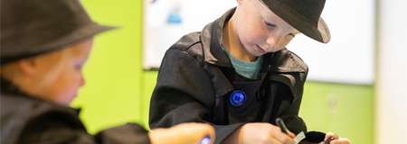 Two children in black detective-style coats and hats examine objects with tools at a table, focusing intently in a brightly lit, green-walled room during a hands-on activity.