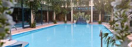 Swimming pool stands still, reflecting ceiling lights; in an indoor atrium bounded by glass walls and columns, surrounded by potted plants and hanging vines, with metal ladders and central steps.