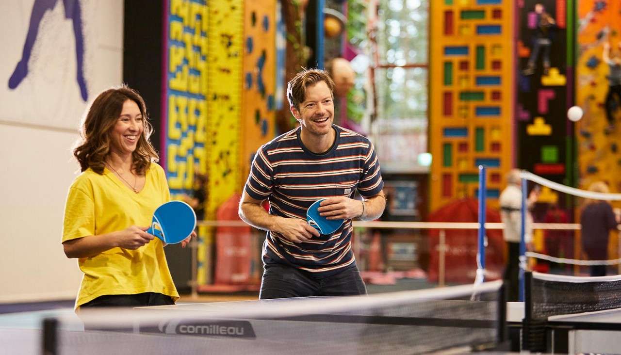 Two adults play table tennis, smiling and holding blue paddles, while standing at a cornilleau table in an indoor recreation center with colorful climbing walls and people in the background.