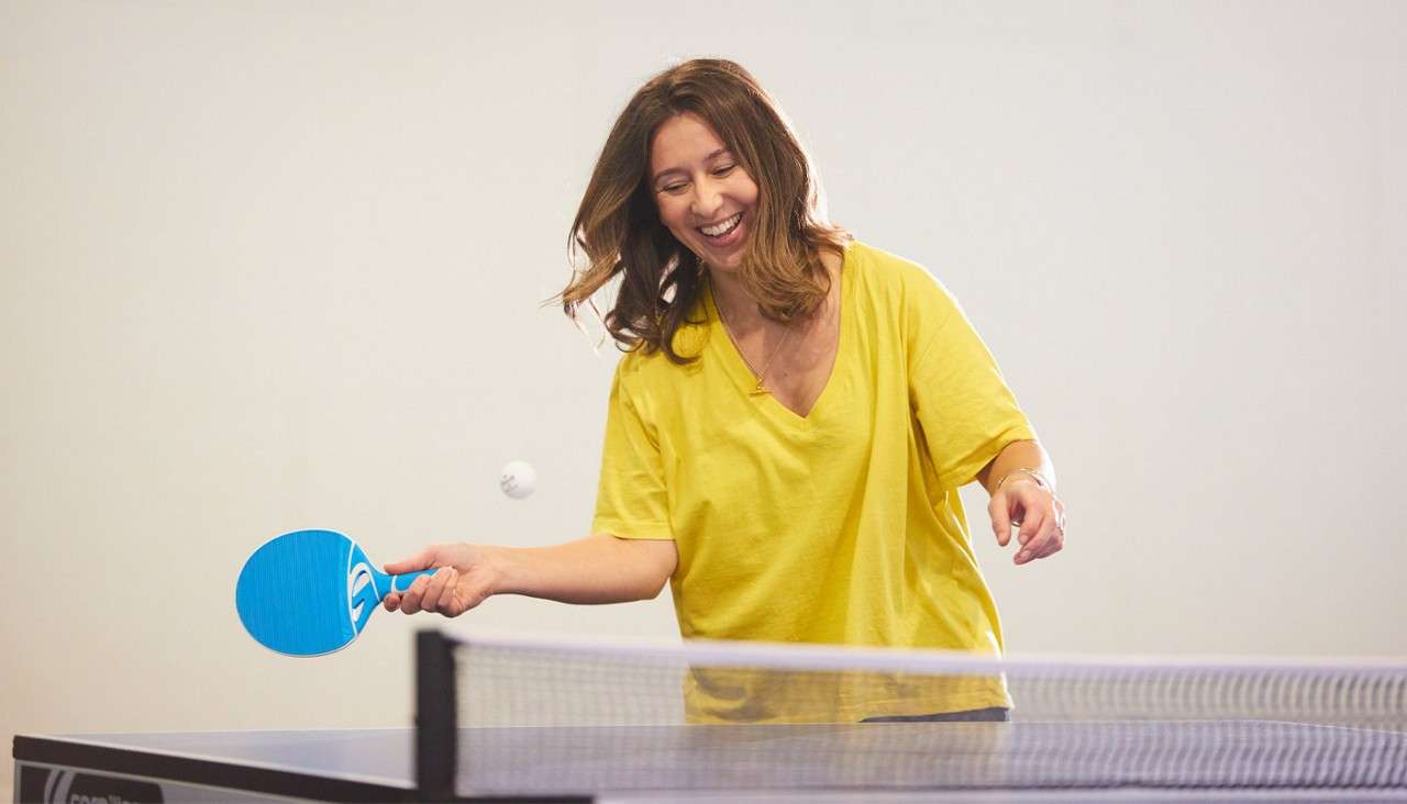 Woman hits a ping-pong ball with a blue paddle, smiling mid-swing; she plays beside a table tennis net in a bright, plain-walled indoor room.