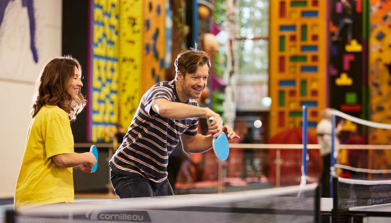 Two people play table tennis, swinging blue paddles at a white ball, in an indoor recreation center with colorful climbing walls. Visible text: “cornilleau” on the table edge.