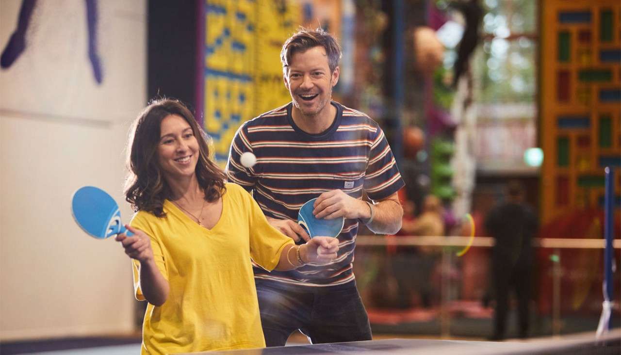 Two adults play table tennis, swinging blue paddles at a white ball, smiling and leaning forward, in a bright indoor recreation center with colorful climbing walls and blurred background figures.