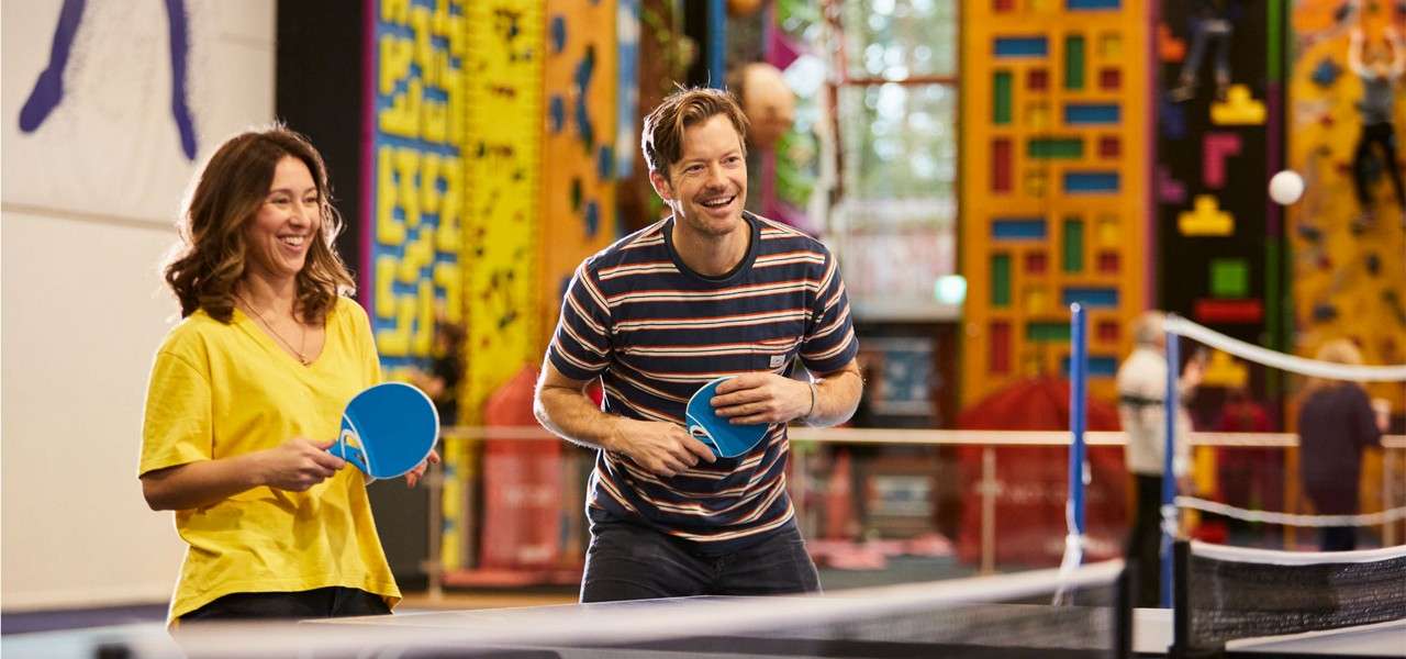 Two adults hold blue paddles, smiling and ready to return a ball, while playing table tennis indoors; behind them, colorful climbing walls, ropes, and people create a lively recreation-center backdrop.