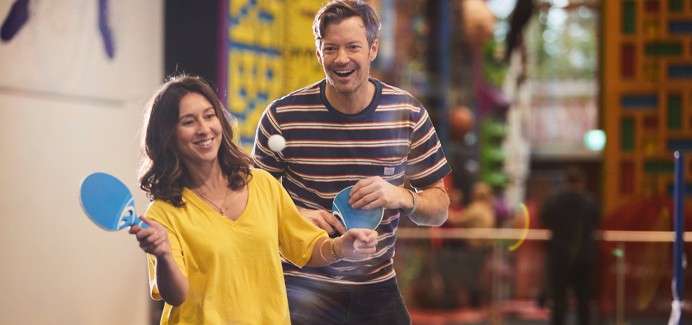 Two adults with blue paddles rally a ping-pong ball, smiling as they move forward in a brightly lit indoor recreation center with colorful climbing structures and a blurred background.
